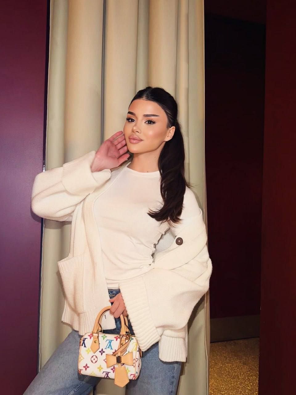 Standing indoors in a cream cardigan and jeans, holding a patterned handbag with a side ponytail.
