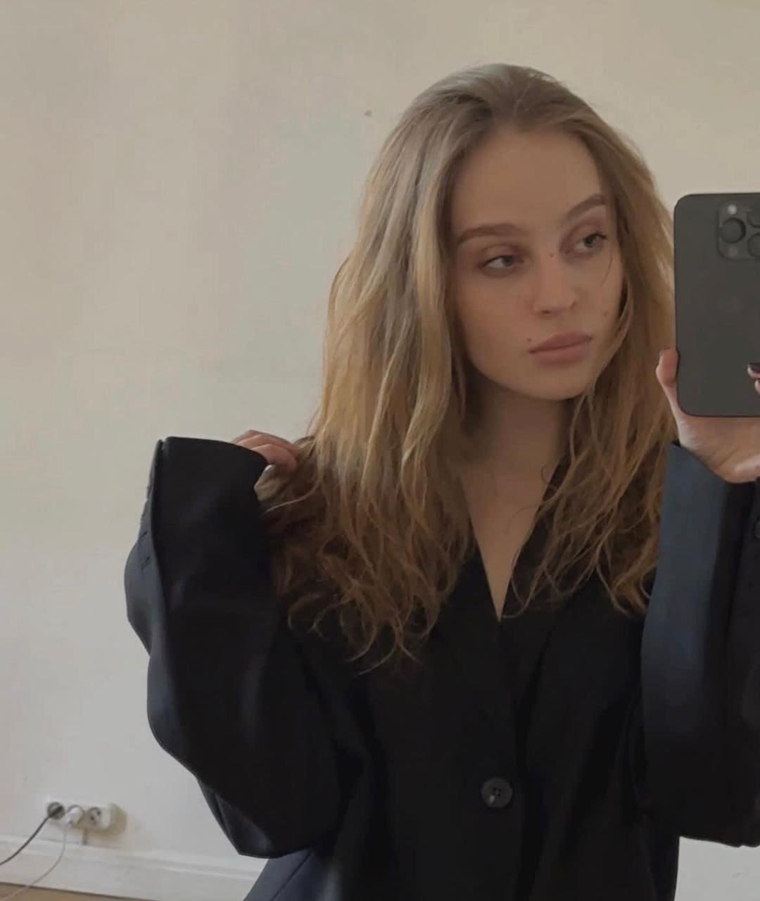 A woman with wavy blonde hair takes a mirror selfie in a black blazer and top, standing before a plain white wall with an outlet.