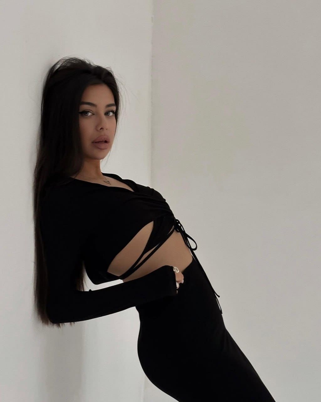 Mirelys leans against a white wall in a black long-sleeved top with side cutouts and a matching skirt, her hand resting on her hip.