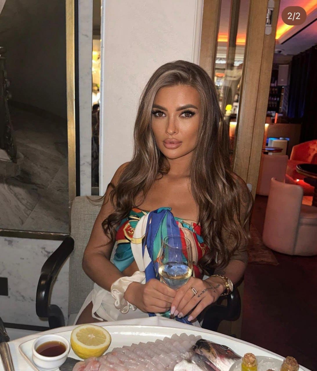 A woman with wavy brown hair sits at a table in a dimly lit room, wearing a colorful strapless top and holding a glass of white wine near a seafood platter.