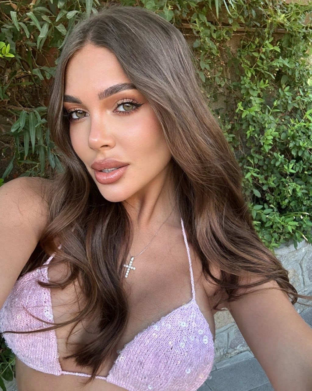 Close-up selfie of a woman in a pink sequined bikini top and cross necklace, framed by green foliage and a stone wall.