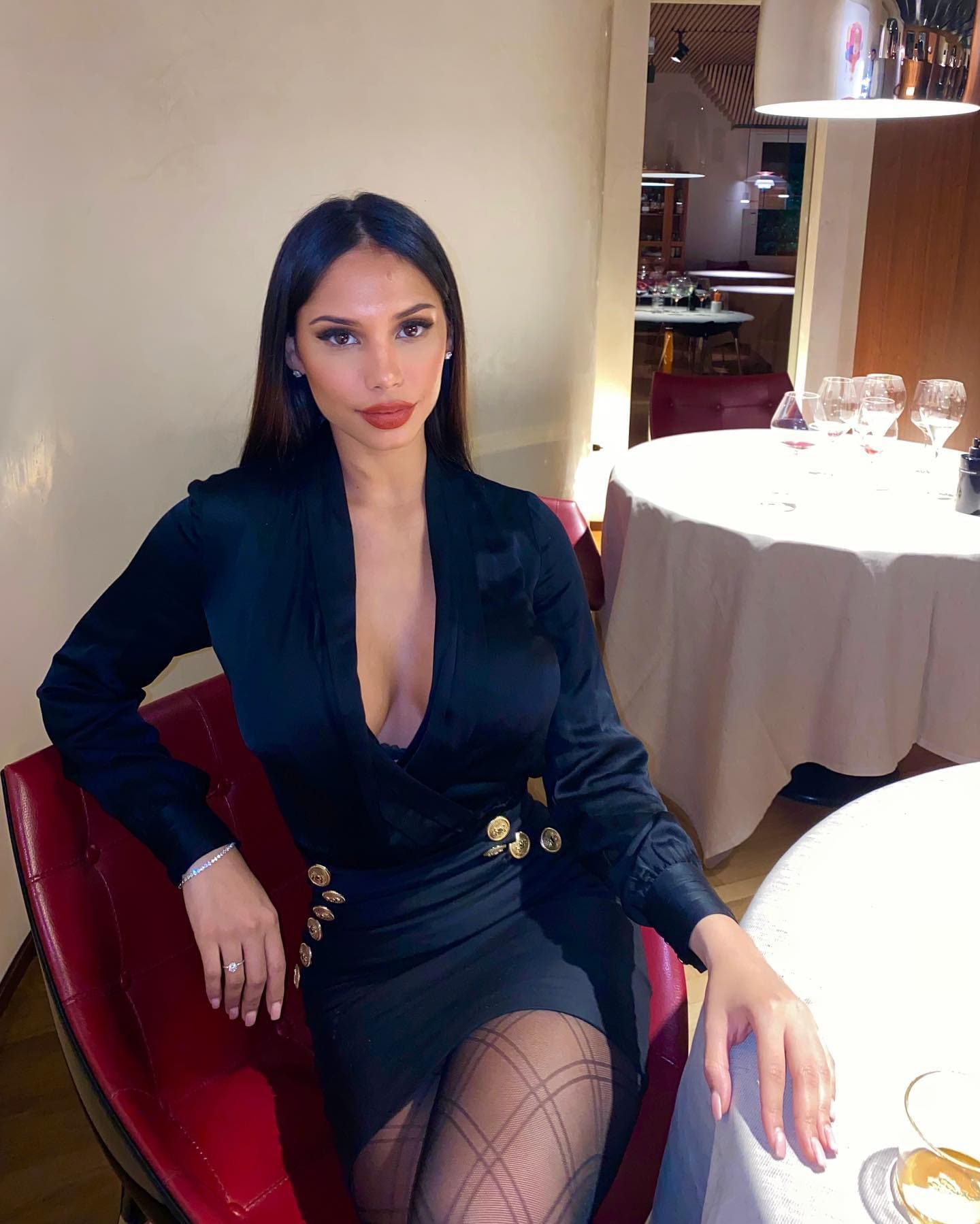 Woman seated in a red chair at a restaurant, wearing a black long-sleeved dress with gold buttons and fishnet stockings.