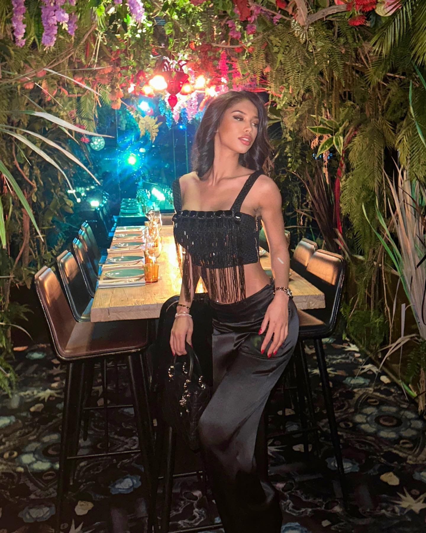 Woman seated on a stool in a plant-filled venue, wearing a black fringed crop top and satin trousers, holding a handbag.