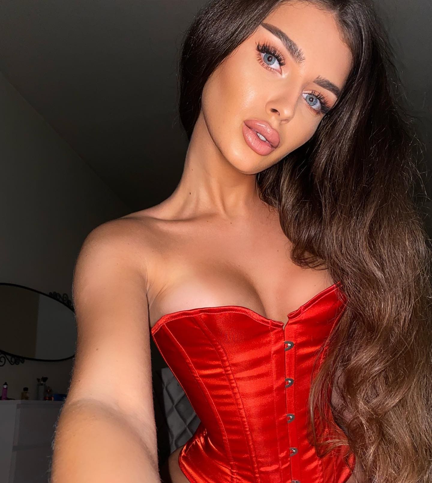 A woman with long dark wavy hair takes a selfie in a red satin corset top, standing in a dimly lit room with a mirror and vanity in the background.