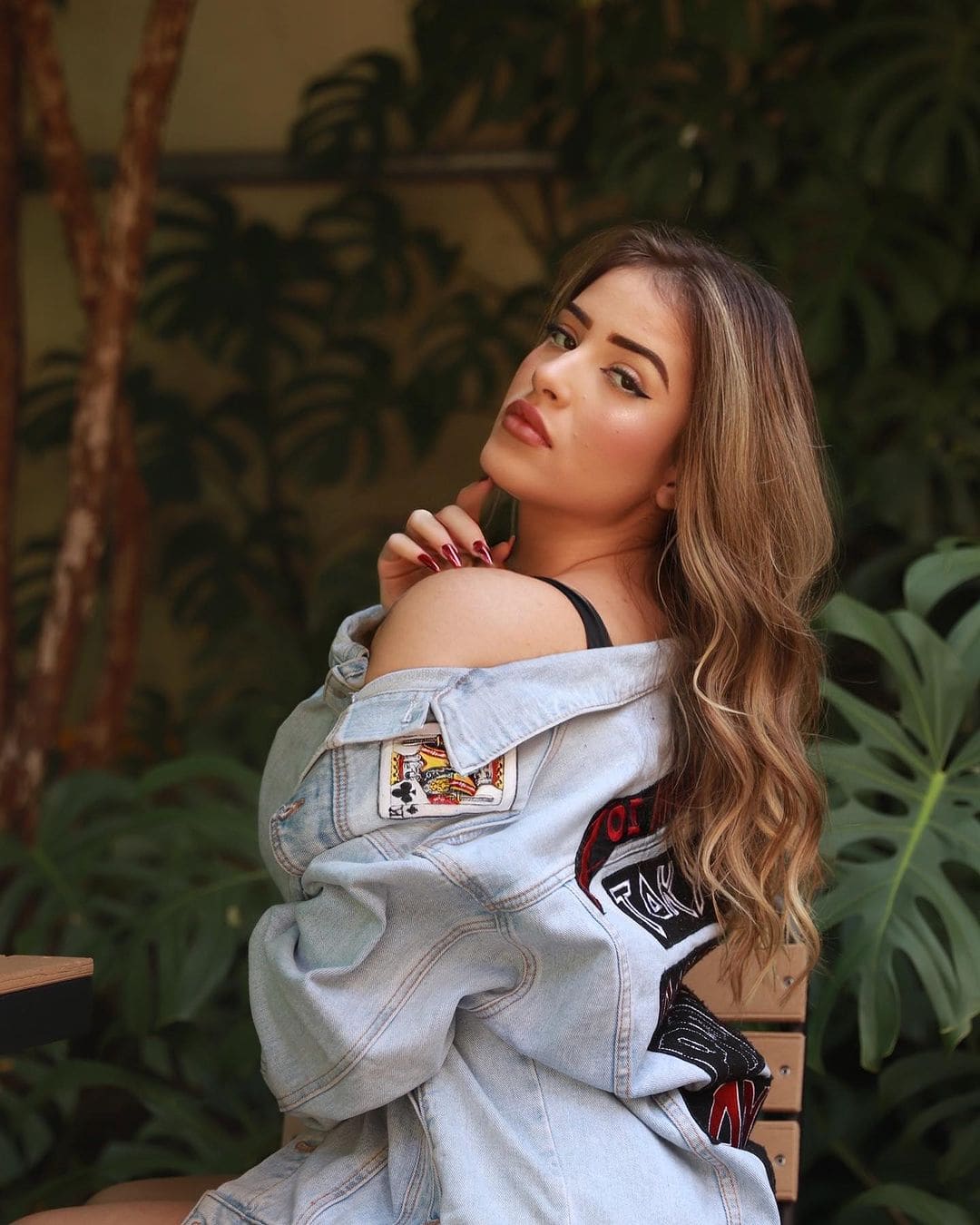 Seated before green foliage, a woman wears a denim jacket with a card patch off one shoulder, hand near her neck.