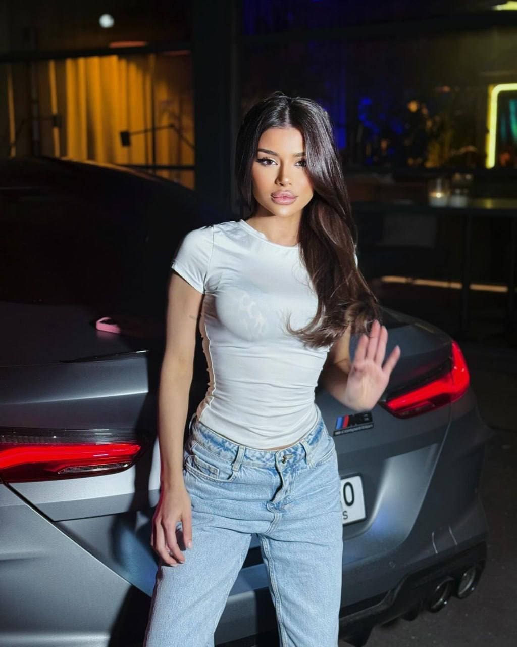 Valentina waves in a white t-shirt and blue jeans, standing before a dark car at night.