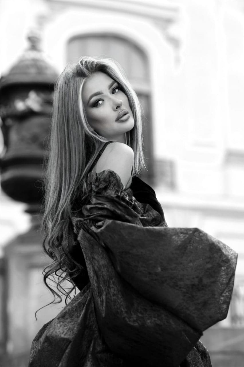 A woman stands in profile wearing a dark off-the-shoulder dress with voluminous sleeves, set against a blurred architectural background with a lamp post.