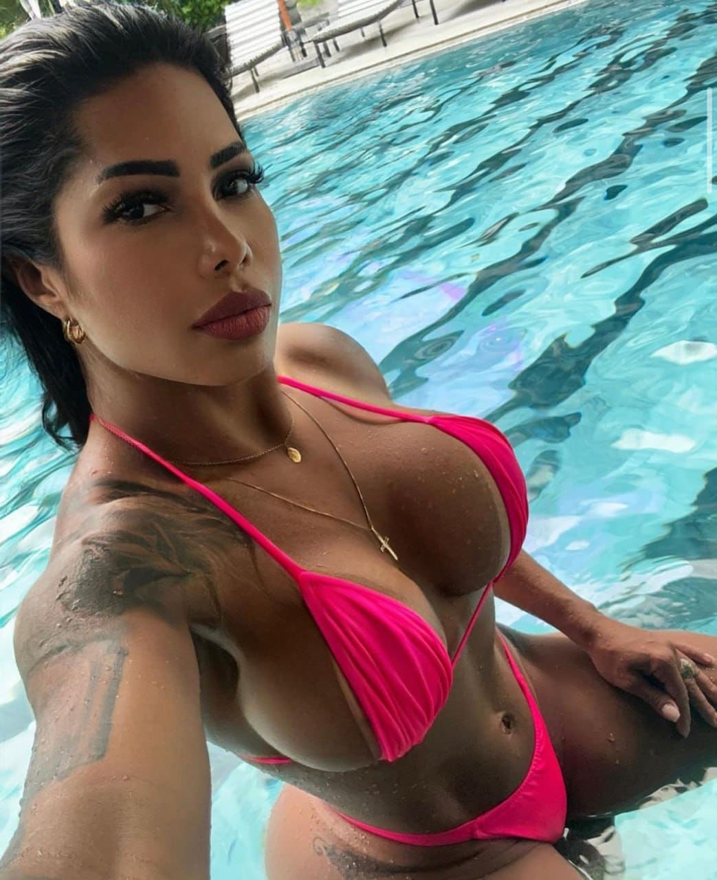 Sitting in a pool, a woman with hair pulled back takes a selfie in a pink bikini, showing tattoos on her right arm and shoulder.