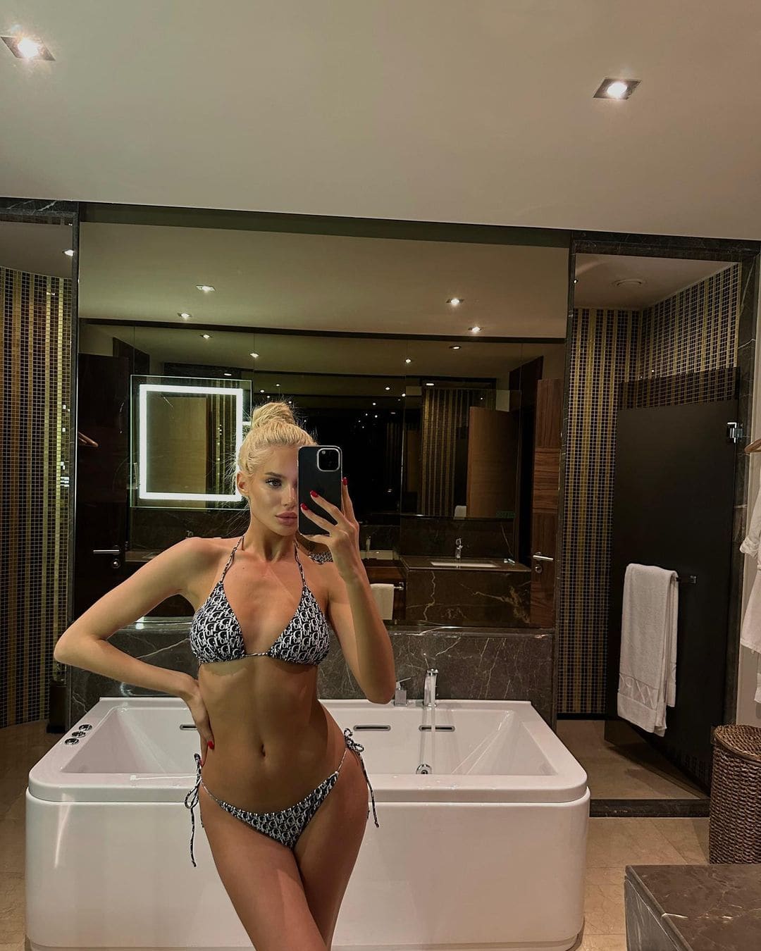 A blonde woman with a bun takes a bathroom mirror selfie in a patterned bikini near a white bathtub.