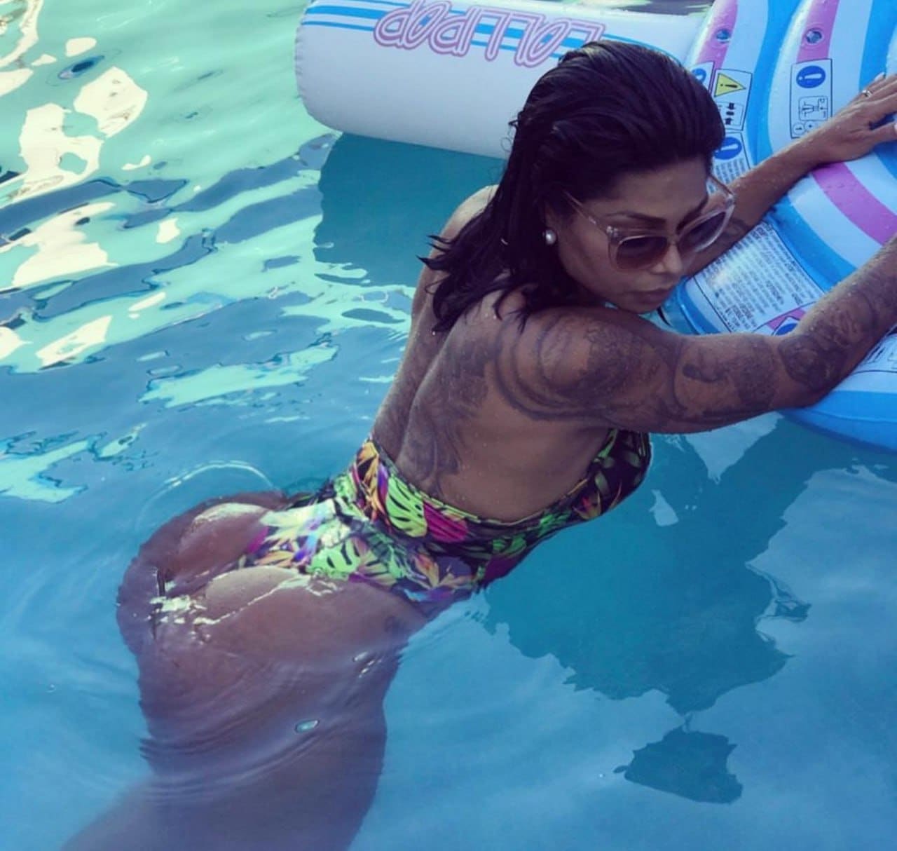 Seen from behind, a woman with wet hair and sunglasses rests her arms on a striped float in a pool, wearing a leaf-patterned swimsuit.
