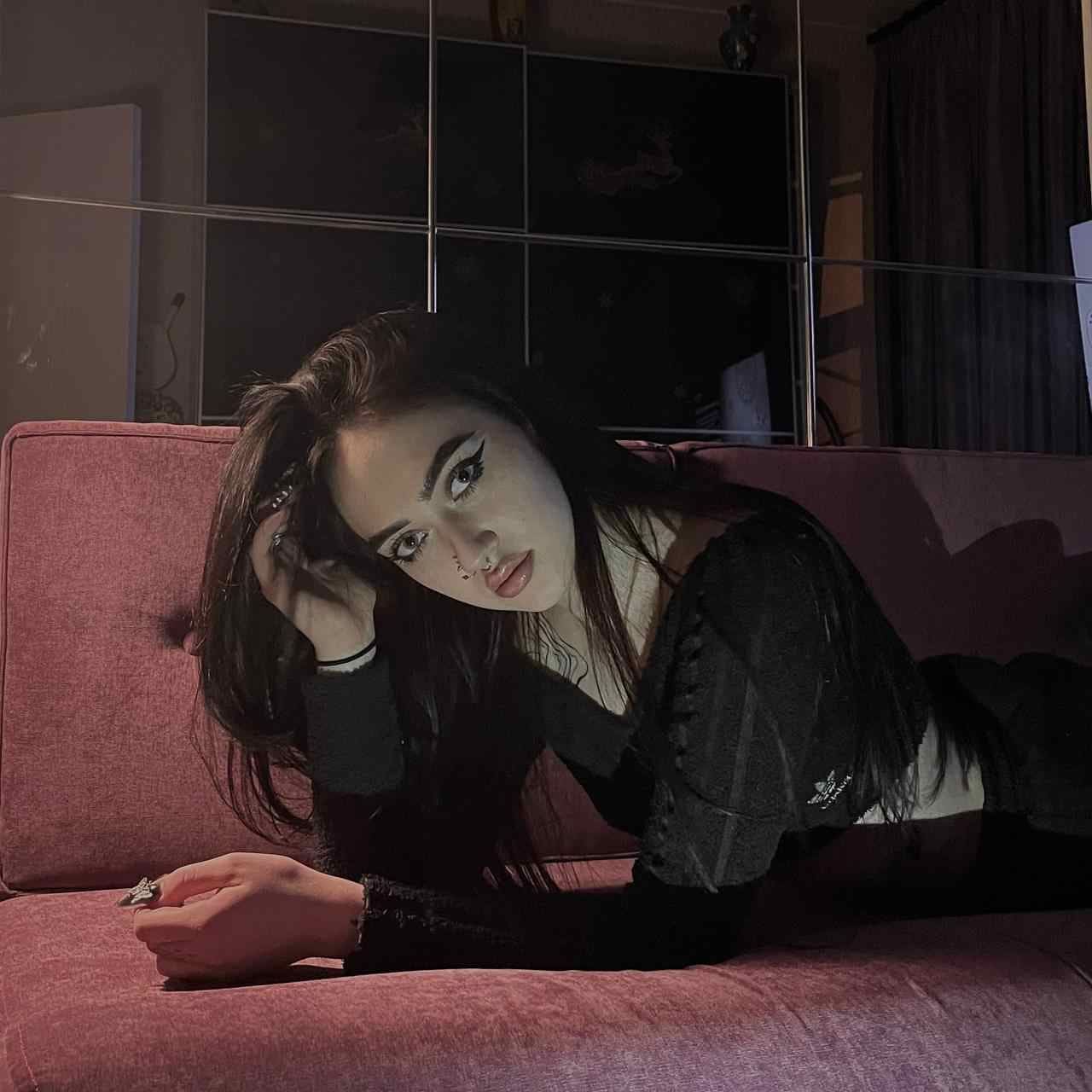 A woman in a black long-sleeved top lies on a pink couch, one hand near her head, in a dimly lit room.