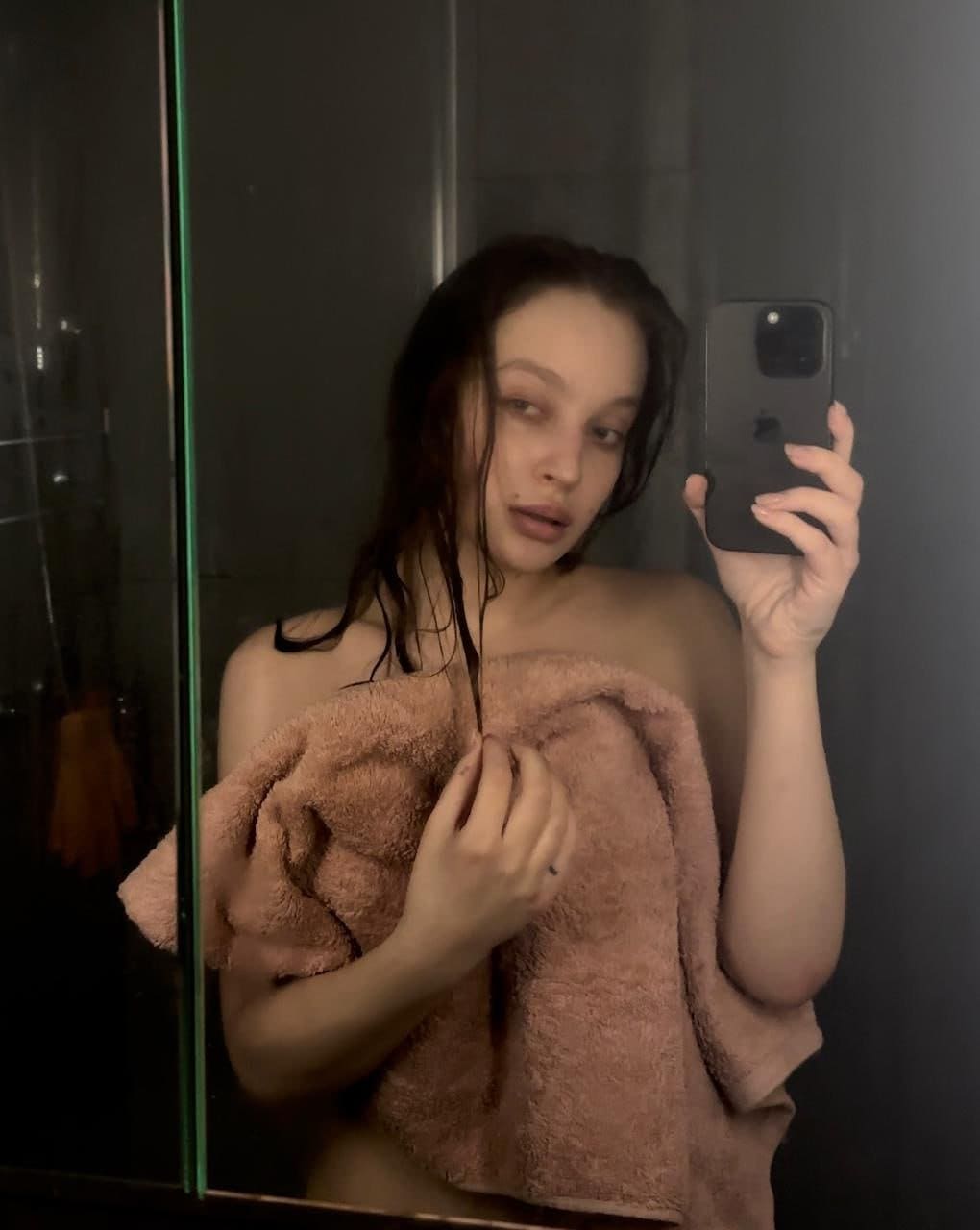 A woman with wet dark hair takes a mirror selfie in a bathroom, holding a phone and a pink towel against a dark tiled wall.