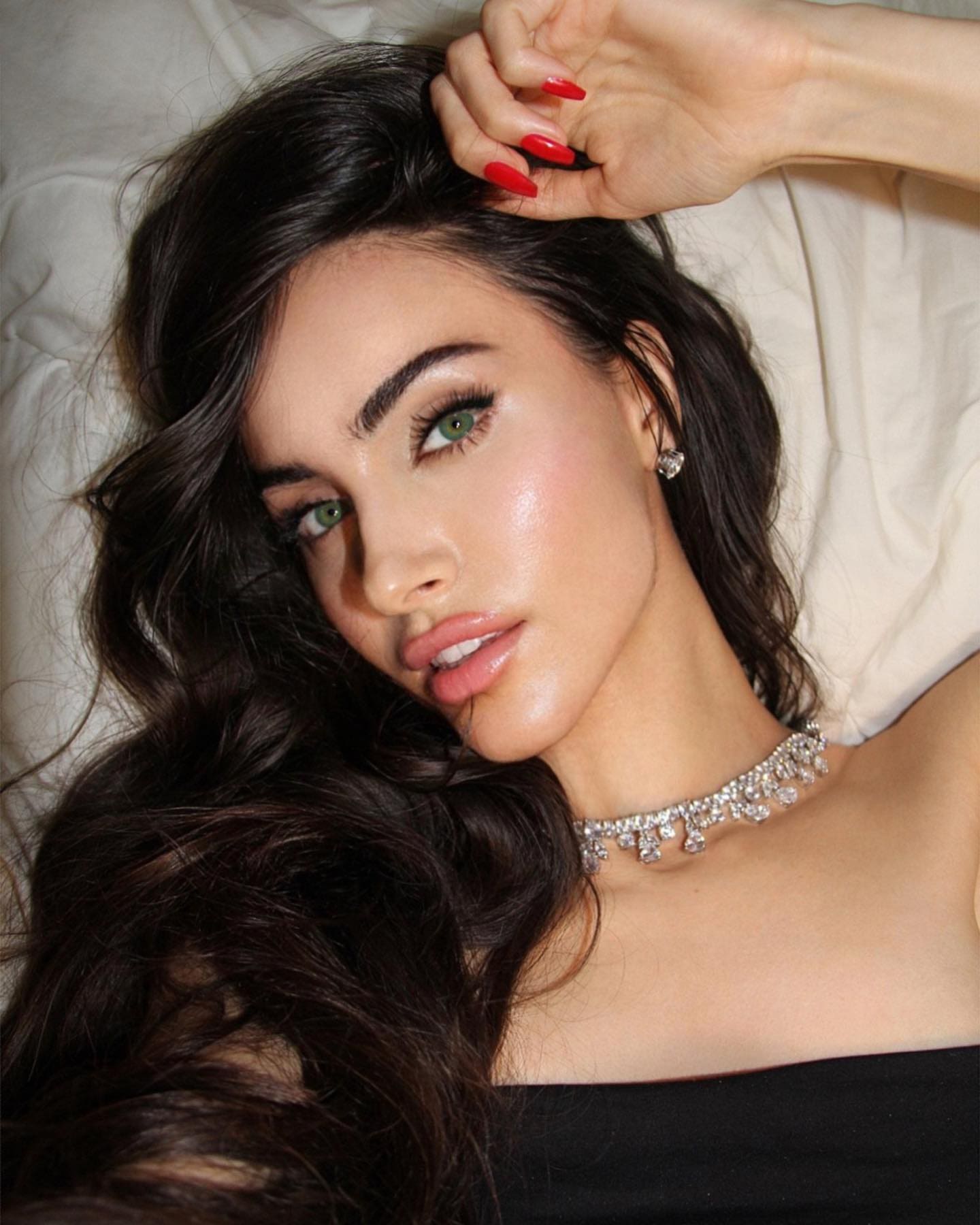 A woman with long dark wavy hair lies on a white bed in a black strapless top and diamond choker, one hand with red nails raised to her head.