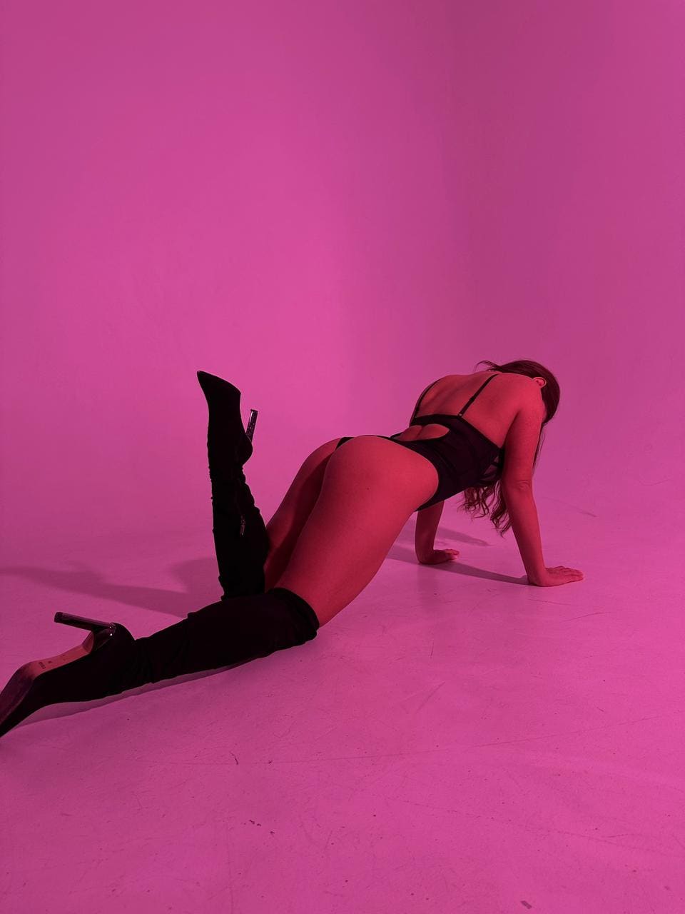 A woman crouches on a pink-lit studio floor in a black strappy bodysuit and thigh-high boots, facing away with one leg raised.
