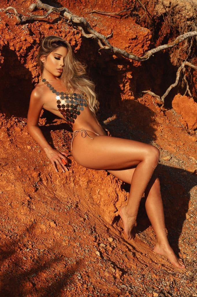 A blonde woman sits on a reddish rocky slope wearing a metallic sequined top and bikini bottom, bare legs visible.