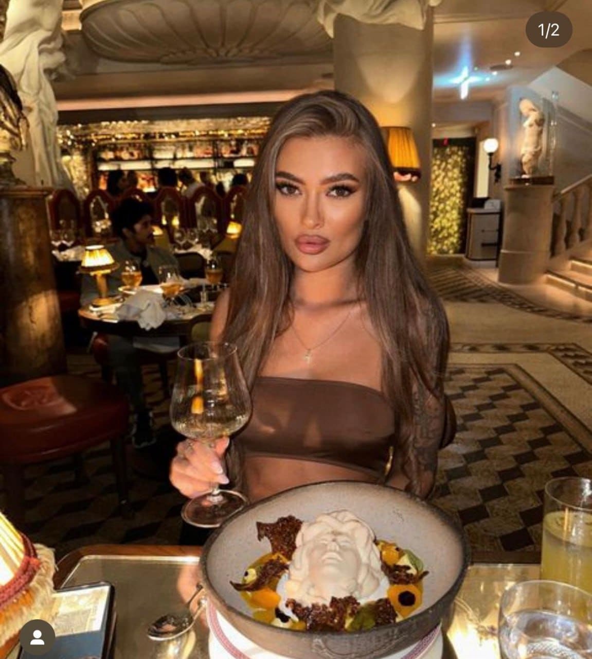 A woman sits at a table in an ornate restaurant wearing a sheer brown strapless top, holding a wine glass with a bowl of dessert in front of her.