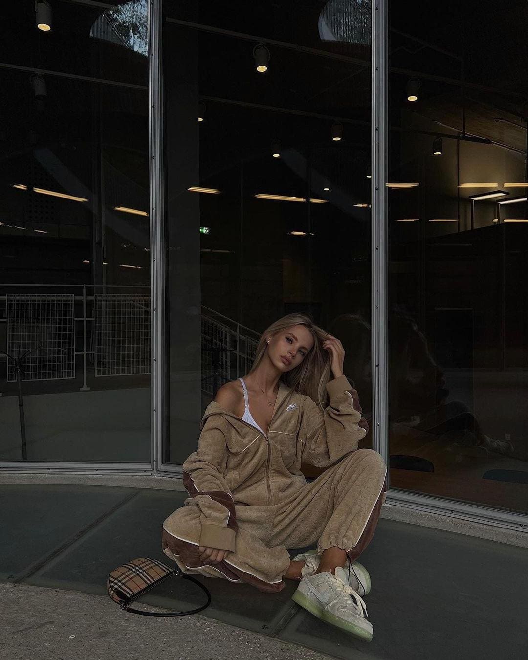 A blonde woman sits cross-legged on a concrete ledge, wearing a tan tracksuit and white sneakers, beside a plaid bag.